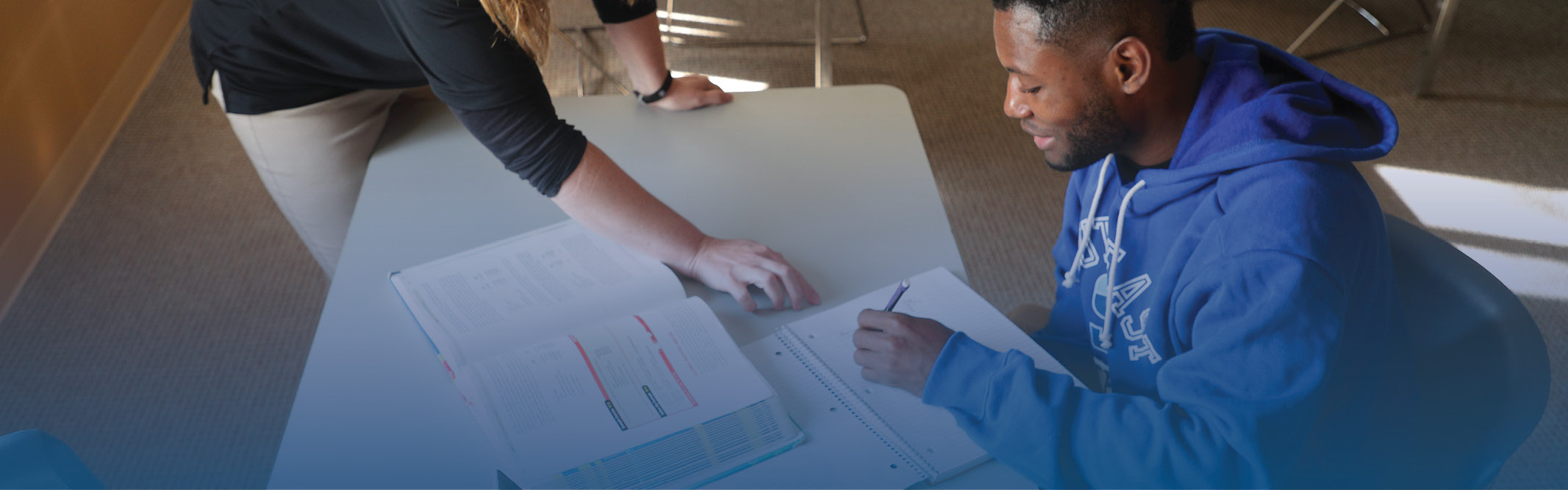 Student at table with open book, writing in notebook with instructor pointing at his page