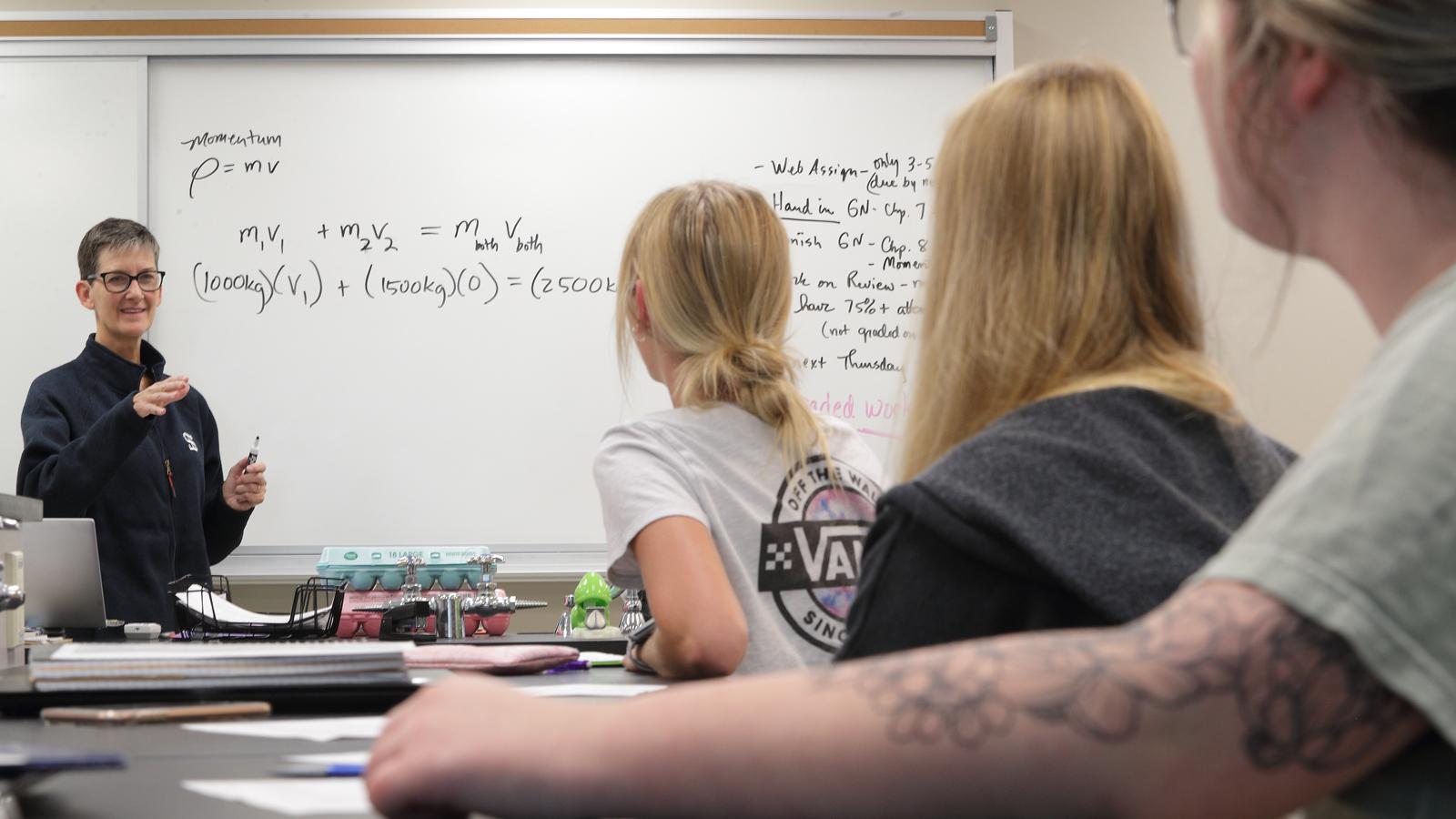 Julie Olson Teaching Physics Julie Olson in front of a whiteboard with equations on it in a physics class