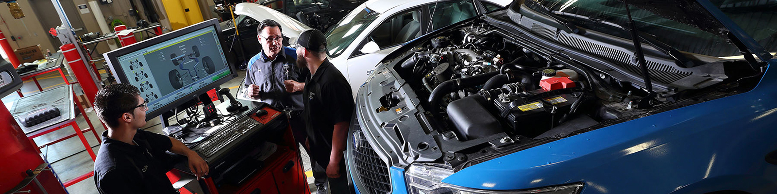 Instructor and students reviewing diagnostics on a blue car with hood up in auto lab