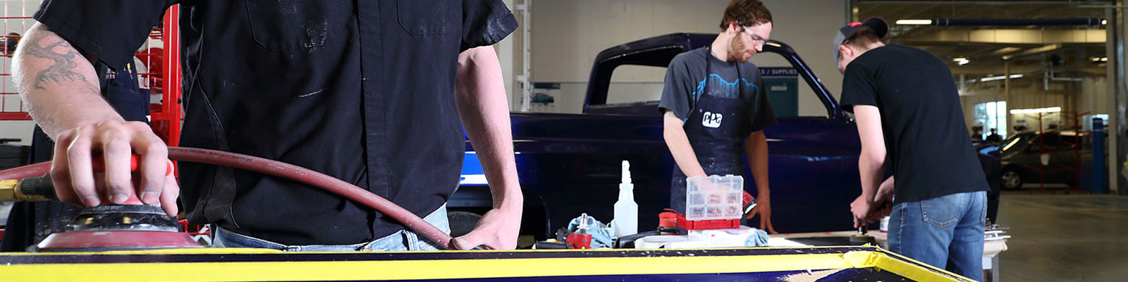 Student in shop shirt buffing tailgate in collision lab, others working in background