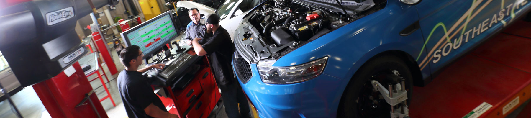 Three men in front of a computer screen next to a blue sedan on a lift with the hood open. 