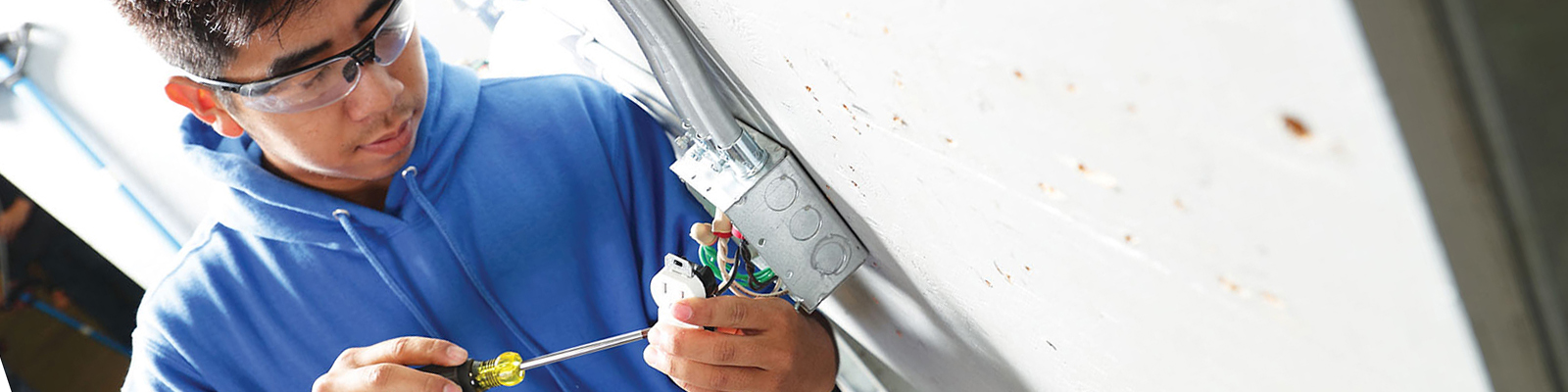 Student wearing safety googles and tool belt installing electrical switch on cement wall