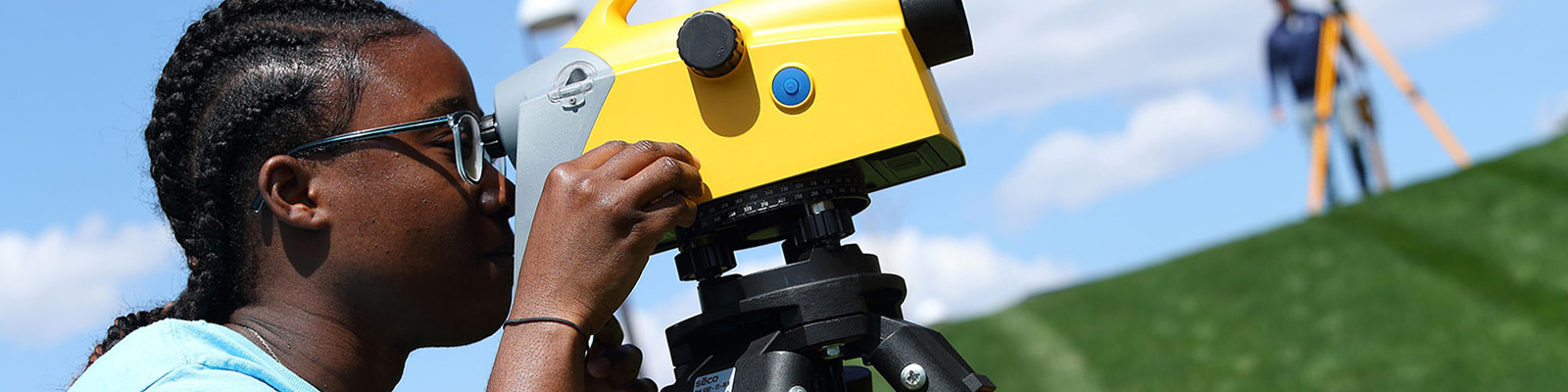 Woman at bottom of grassy hill, looking into surveying equipment. Soft focus background on another student.