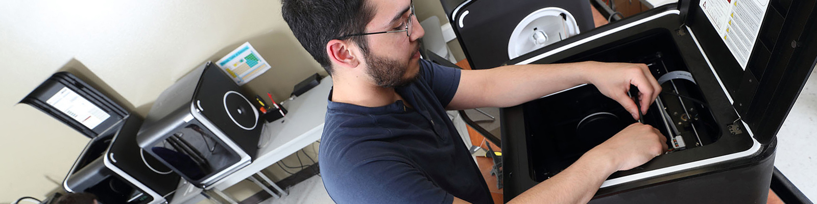 Male student in front of 3D printer, making adjustments in room full of 3D printers.