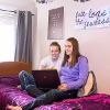 Two students sit on a bed in student housing looking at a laptop. 